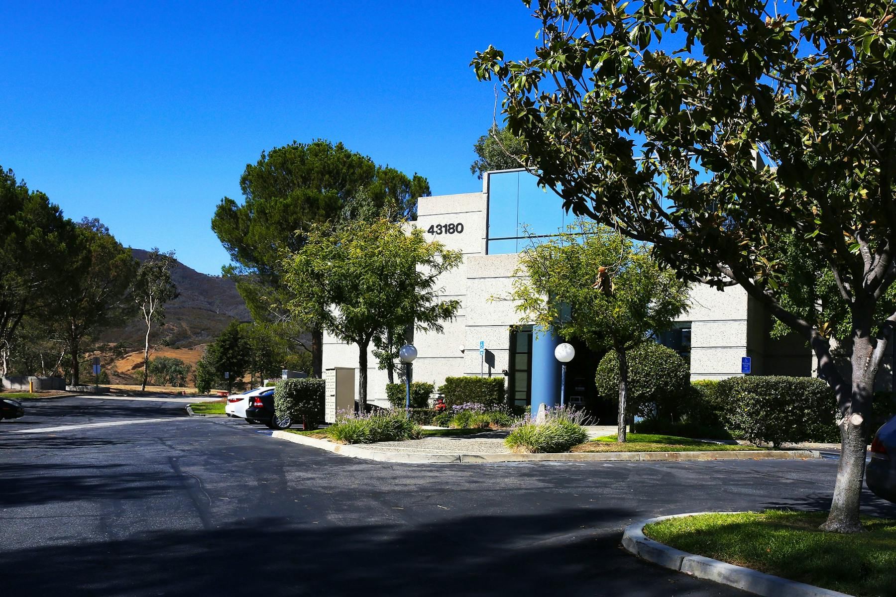Two-story light-grey office building with the address numerals 43180, a blue entry column, mature trees and parking in Temecula